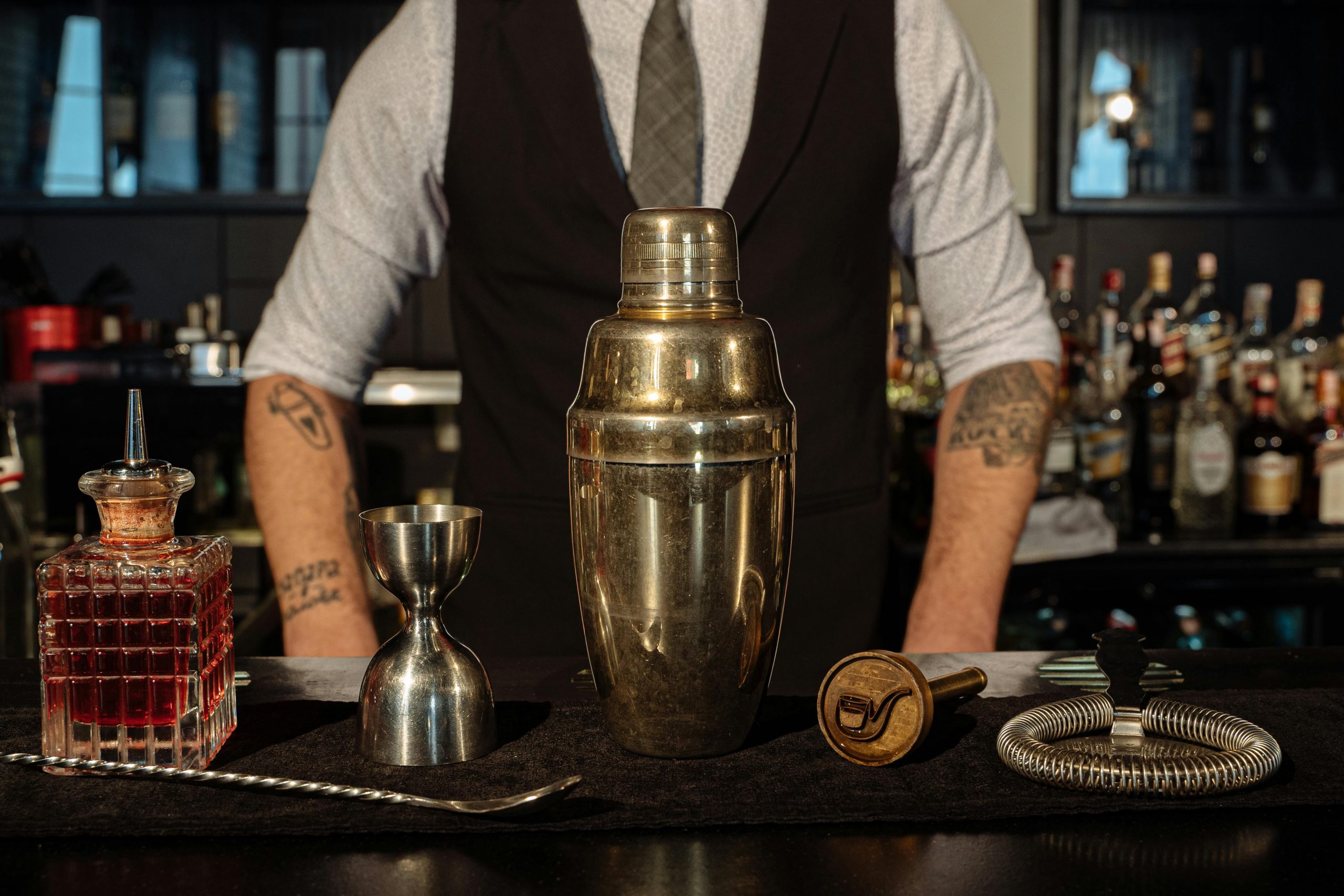 bartender standing behind a counter with a stainless steel shaker, measuring cup, and bottle of liquor